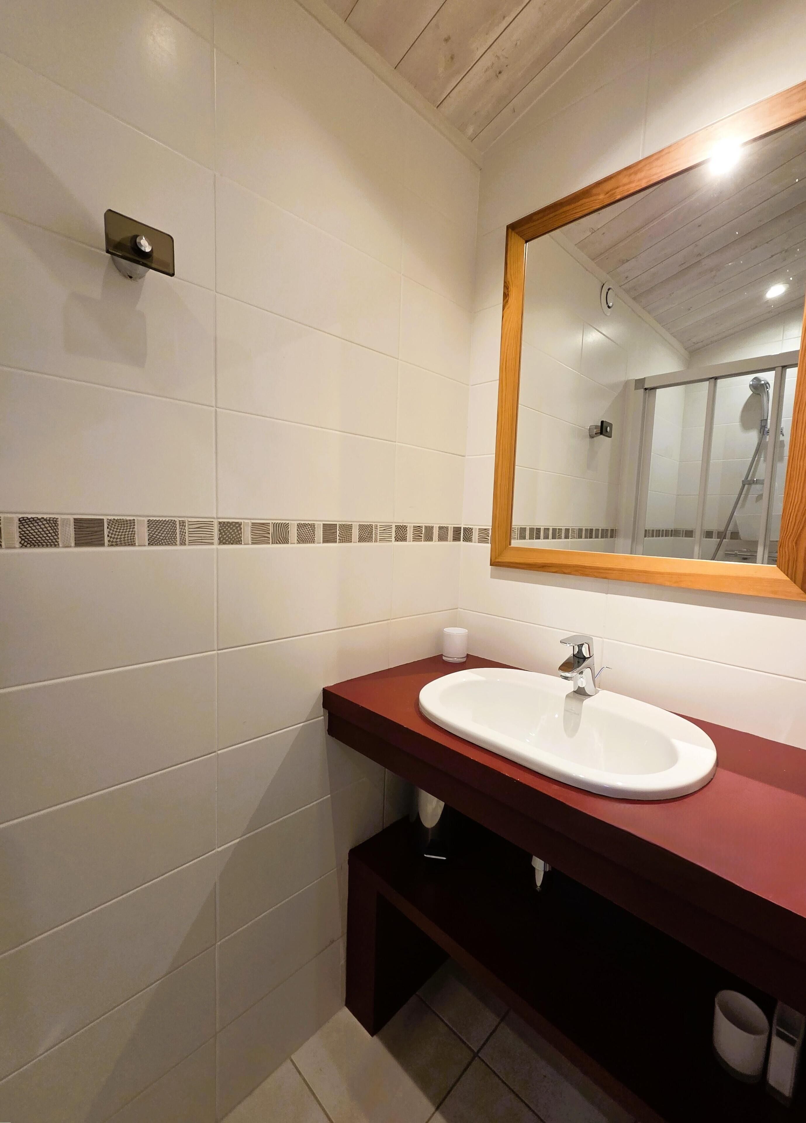 Une salle de bain avec des murs recouverts de carreaux blancs, un plafond en bois, un grand miroir et un lavabo blanc doté d’un plan de travail rouge.