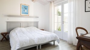 Un dormitorio con cama, cabecera de madera, mesita de noche, una silla, ventana con cortinas e imagen en la pared.
