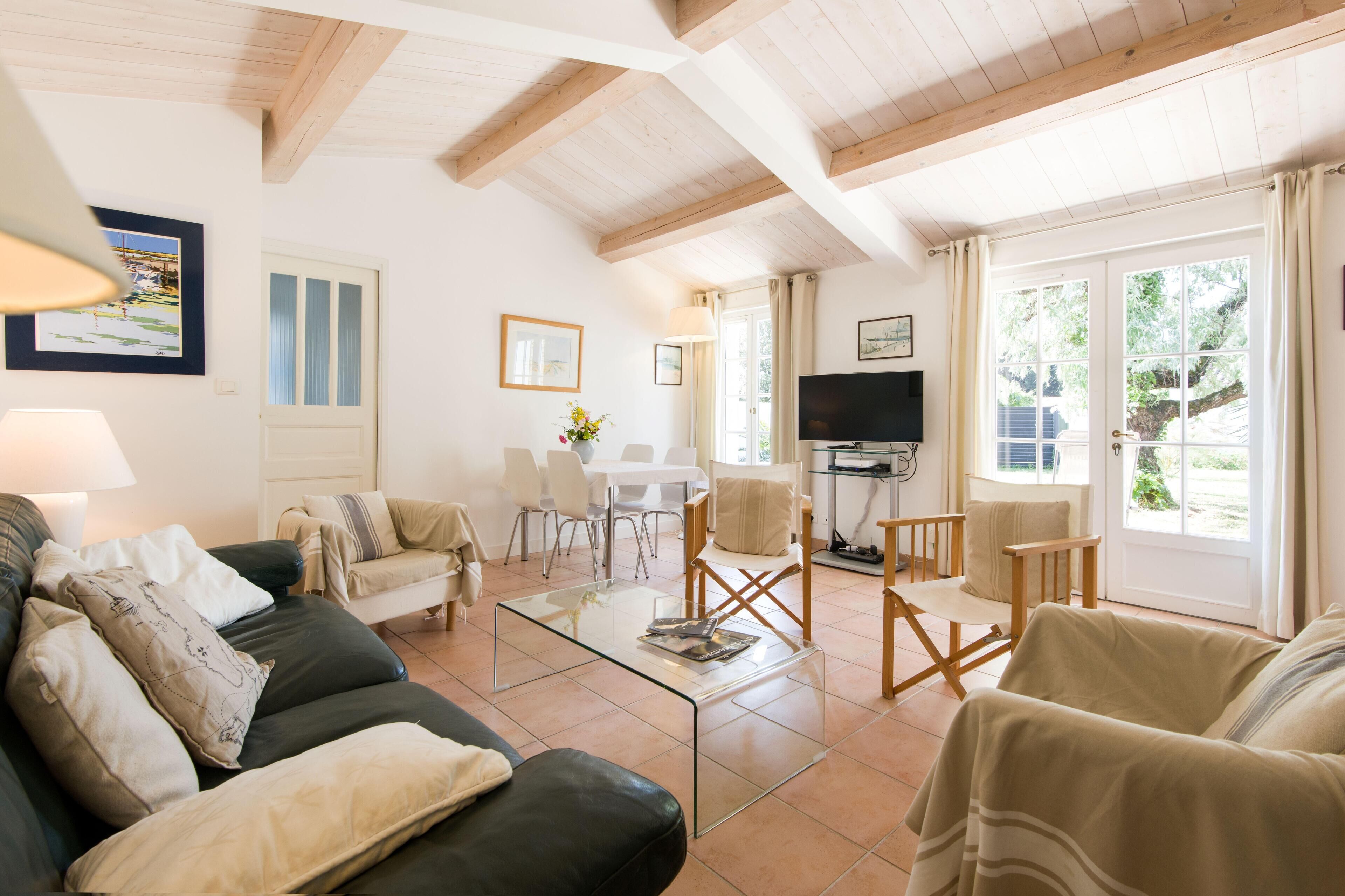A spacious living room with a glass coffee table, leather sofa, and wooden chairs. There is a TV, a dining table, and a door leading outside.