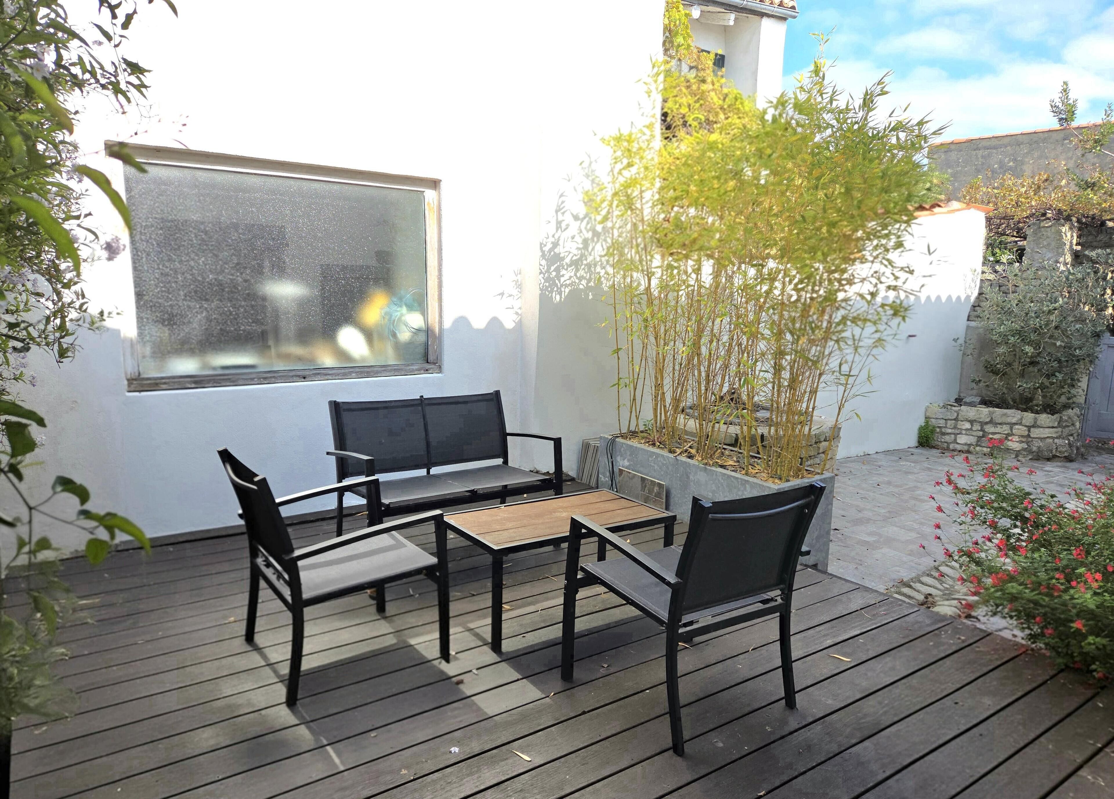 Una terraza de madera con una mesa y sillas, una ventana y un jardín con plantas y flores.