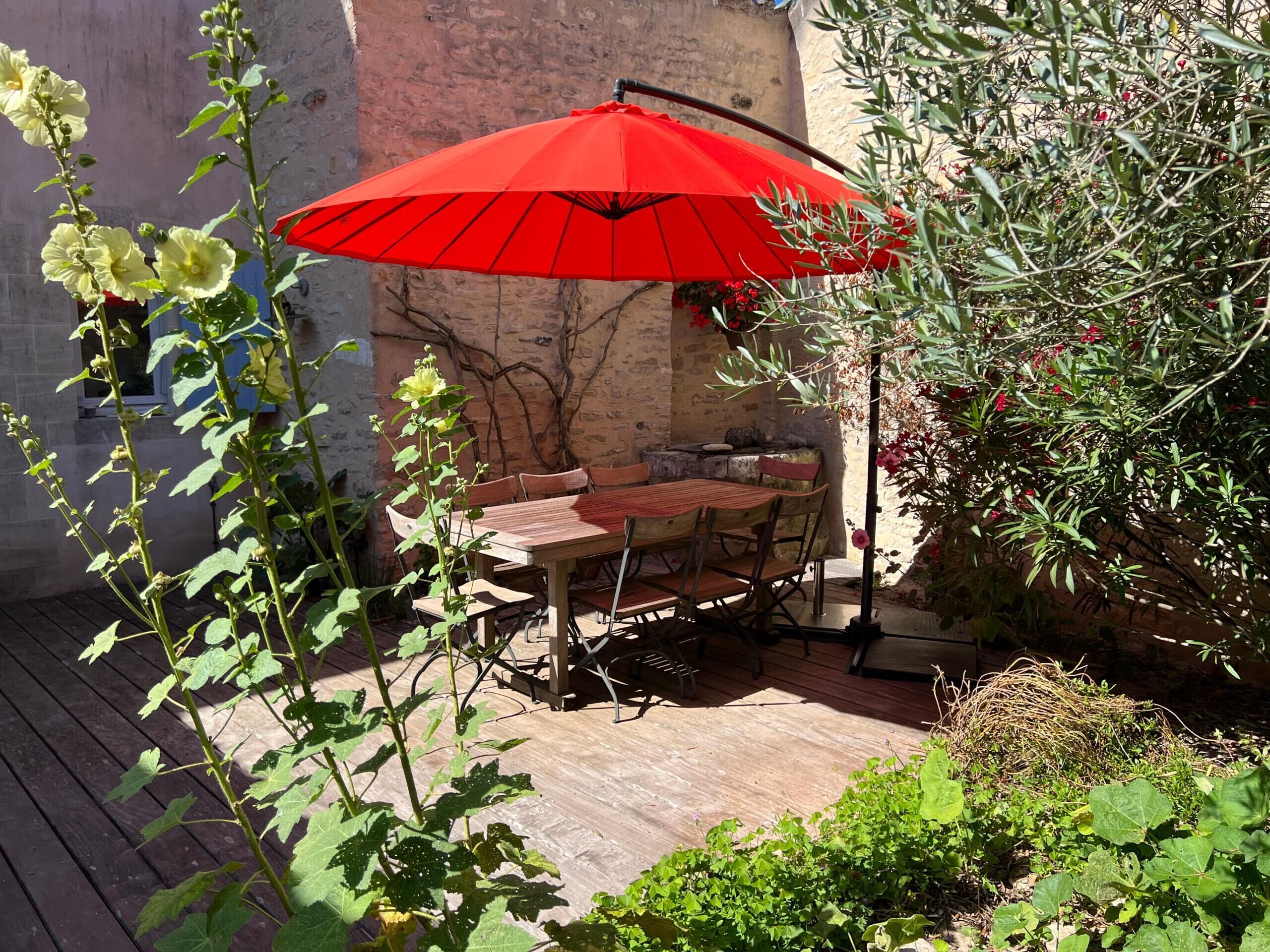 An outdoor patio with a wooden table and chairs, a red umbrella, and various plants.