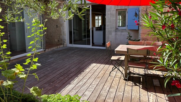 Una terraza de madera con un paraguas rojo, una mesa con sillas y un ventanal corredero que da a una cocina.