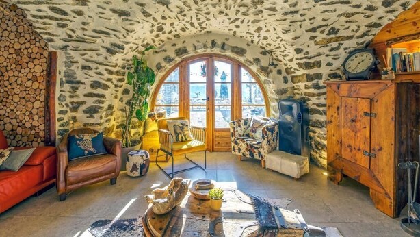 A rustic interior with stone walls, a wooden arched window, a red sofa, and a wooden cabinet.