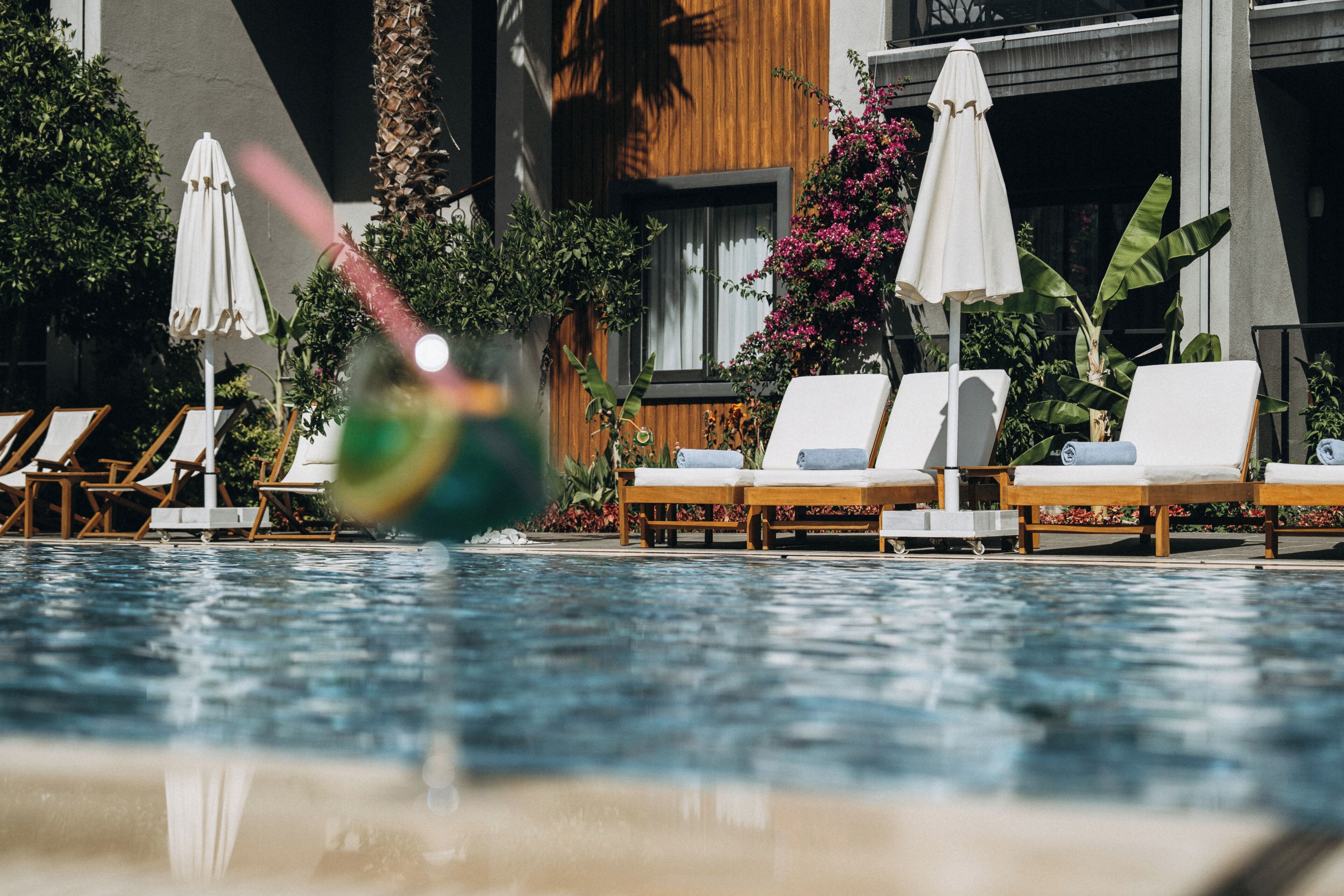A pool with lounge chairs and umbrellas, surrounded by plants and a building with wooden paneling.