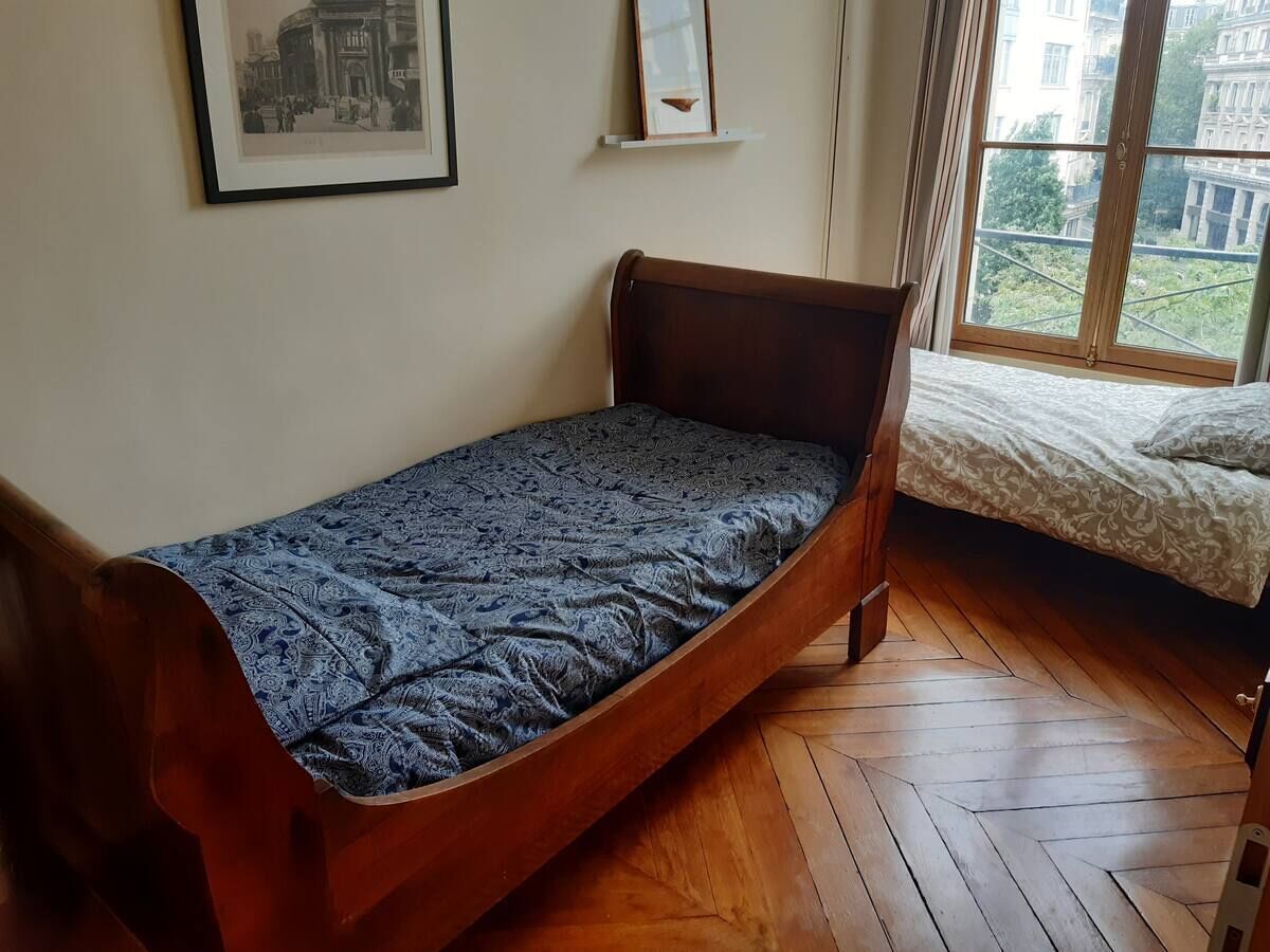 A single bed with a patterned bedspread in a room with wooden flooring and a window with a view of buildings.