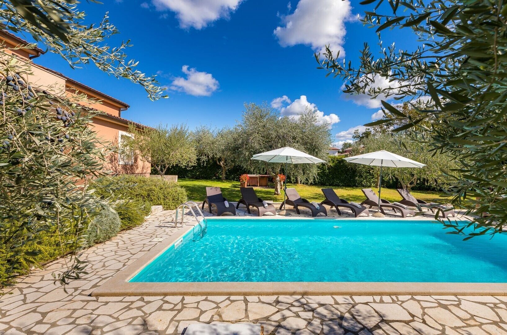 Une piscine entourée de verdure et dotée d’une allée en pierre, avec des chaises longues et des parasols à proximité.