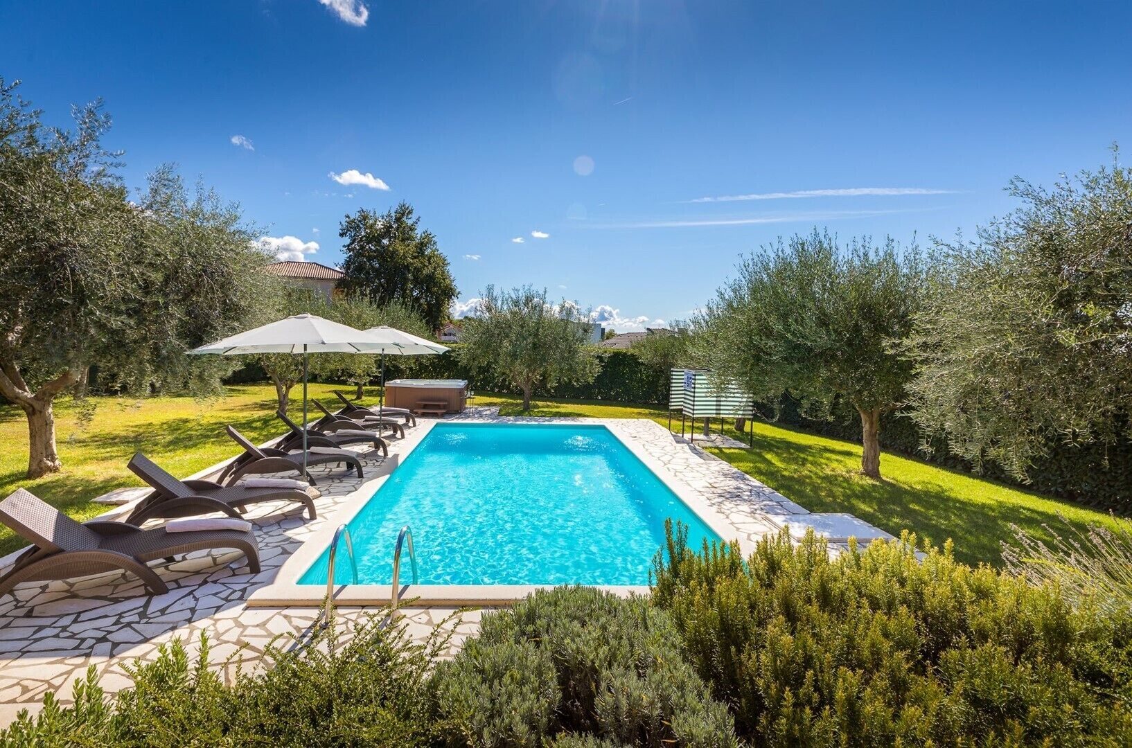 A rectangular swimming pool with a tiled deck, surrounded by lounge chairs and umbrellas, set in a lush garden with trees and shrubs.