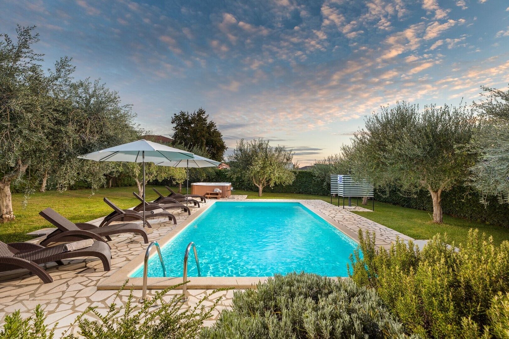 A rectangular swimming pool with a tiled deck, surrounded by lounge chairs and an umbrella.