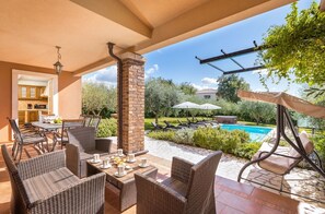 Eine überdachte Terrasse mit Korbmöbeln, einem Pool und Blick auf den Garten.