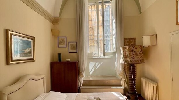 Un dormitorio con cama, ventana con cortinas, radiador y un cuadro en la pared.