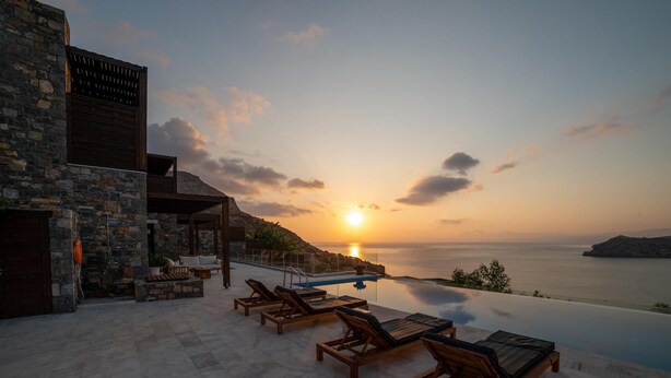Vista al atardecer desde una piscina infinita con sillones de descanso, un edificio de piedra y cielo despejado.