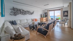 Un salón espacioso con un sofá grande, una mesa de centro y un mueble para televisión. Hay un balcón con vistas al mar.