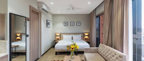 A hotel room with a bed, a sofa, a coffee table with a book and flowers, a mirror, and a window with curtains.