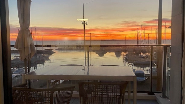 Ein Balkon mit Tisch und Stühlen, der auf einen Yachthafen mit Booten und einem Sonnenuntergang blickt.