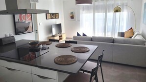 Eine moderne Küche mit einer zentralen Kochinsel, einem Esstisch und einem grauen Sofa in einem hellen Wohnbereich mit großem Fenster und Vorhängen.