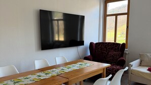 A dining area with a long wooden table, white chairs, a flat-screen TV mounted on the wall, and a window with a view of a green landscape. - Lilienstein (Königstein)