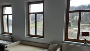 A room with a sofa, a window with a view of a river and buildings, and a radiator. - Lilienstein (Königstein)
