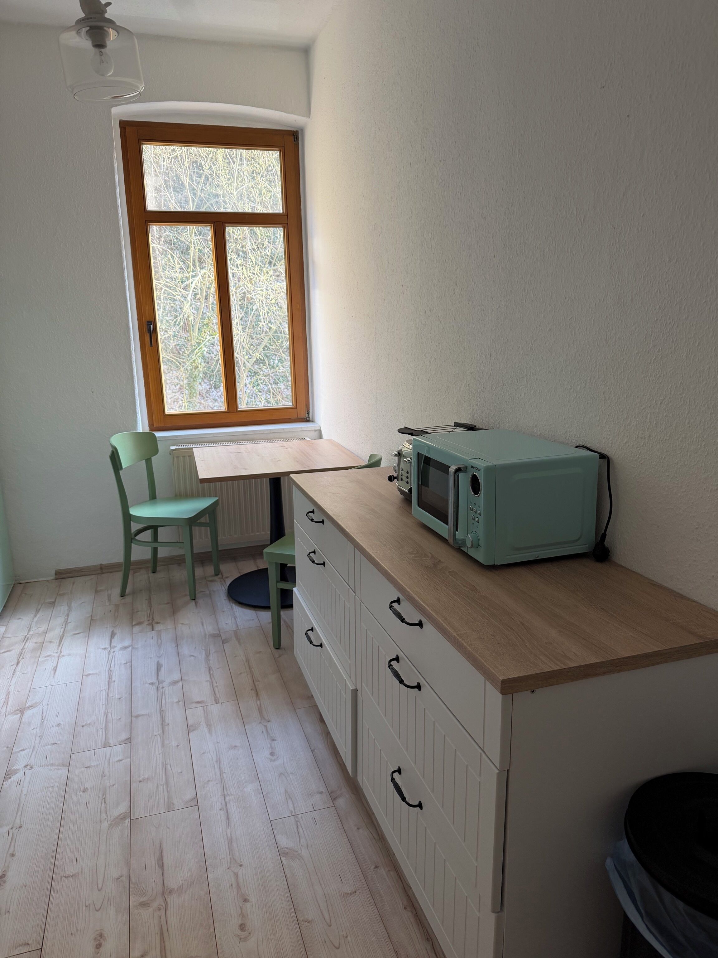 Una cucina con una finestra, un forno a microonde, una sedia e un termosifone.
