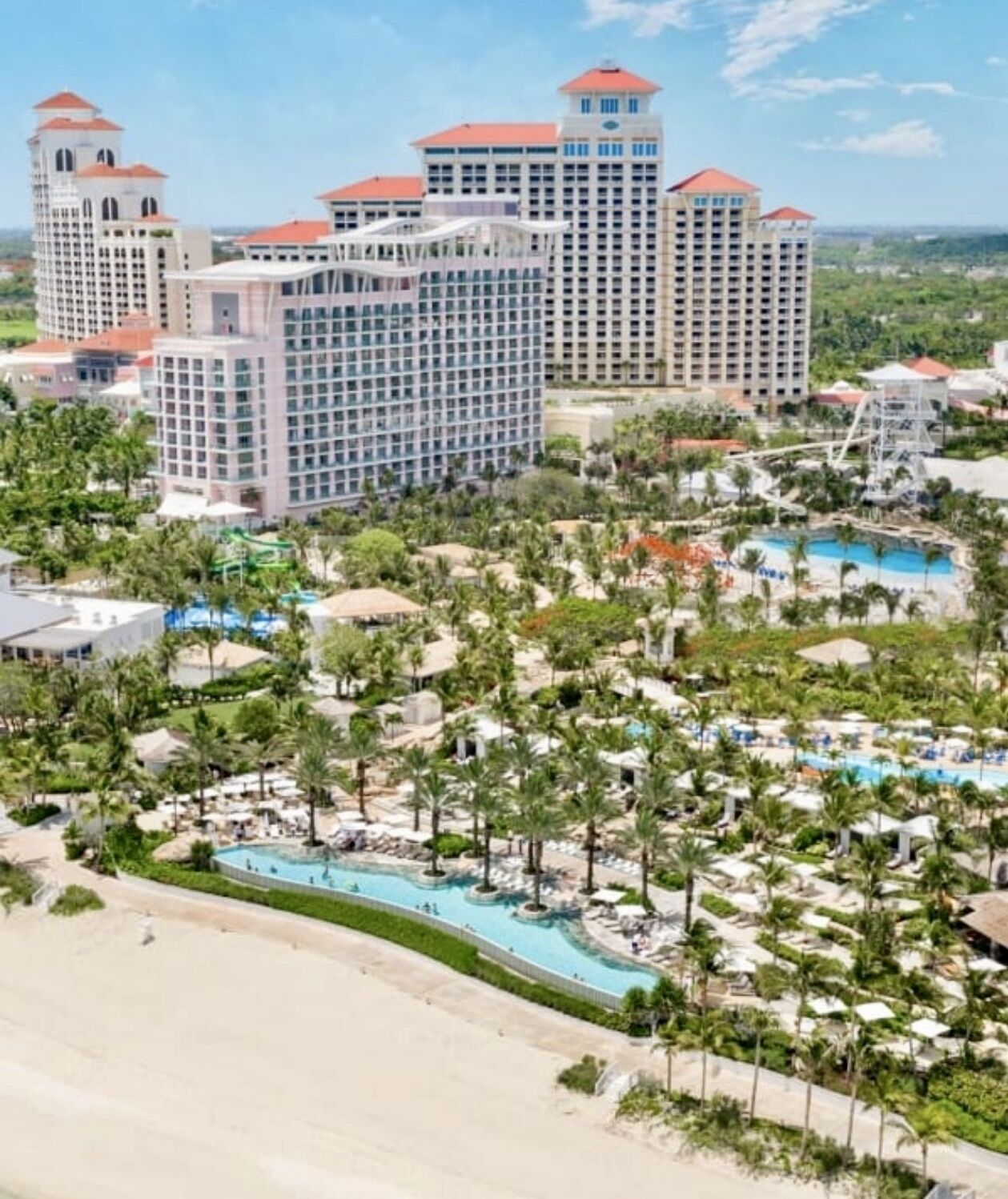 A resort with multiple high-rise buildings, a central pool area surrounded by palm trees, and a beach in the foreground.