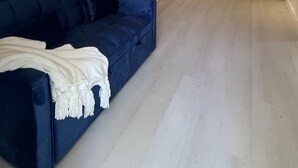 Een ruime woonkamer met een blauwe bank, een keuken met witte kasten en een houten plafond.