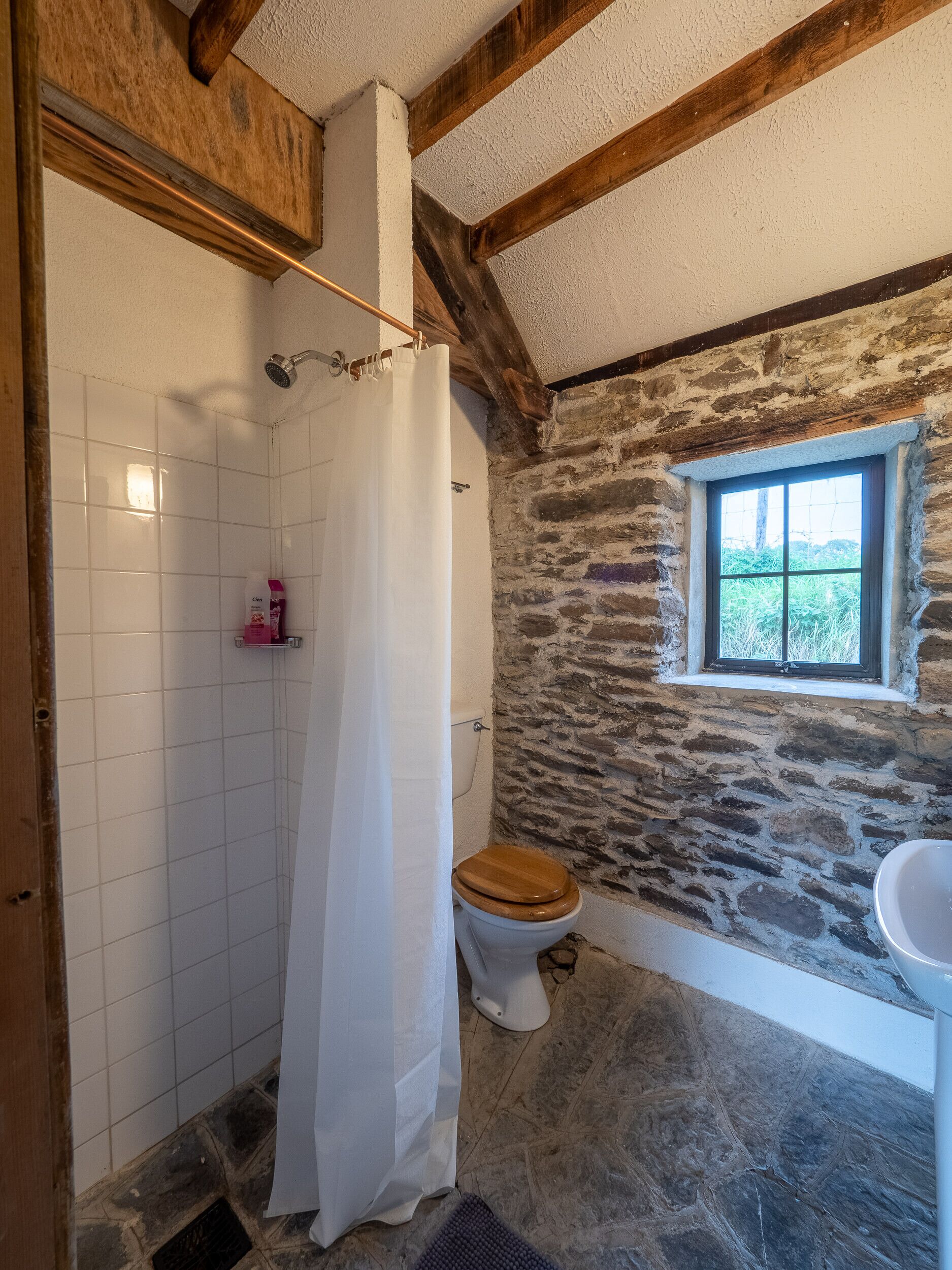 Un baño con pared de piedra, techo de madera, inodoro blanco y ventana.