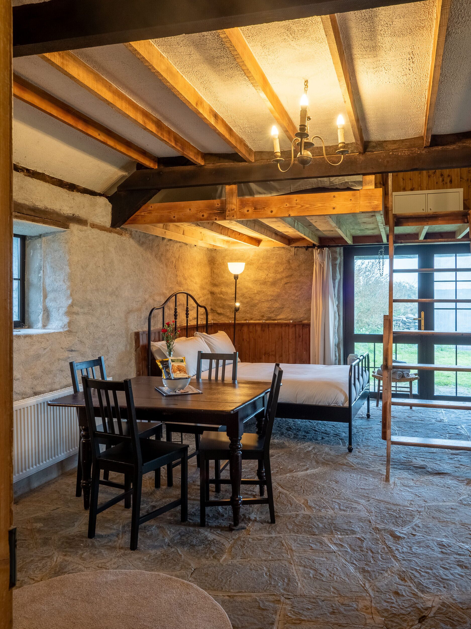 A rustic room with a stone floor, wooden beams, a bed, a table with chairs, and a window with curtains.