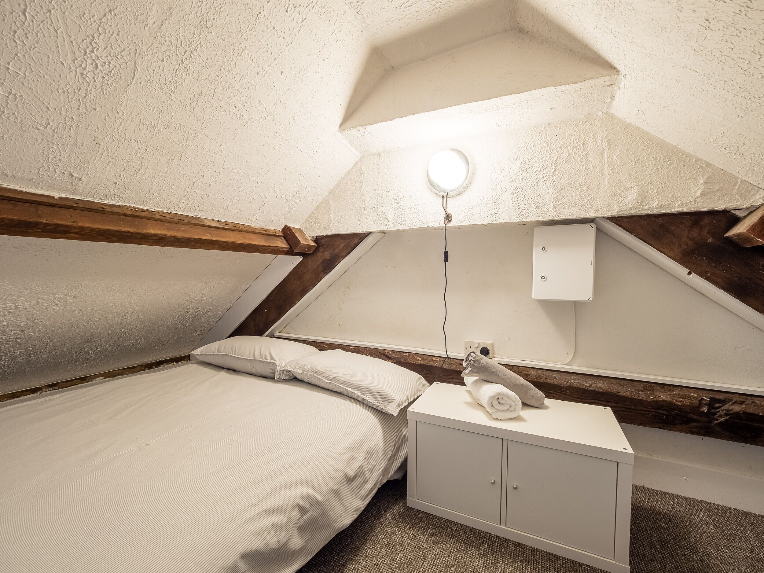 Une petite chambre mansardée, chaleureuse, avec un plafond en pente, un lit simple, une table de chevet et un luminaire fixé au mur.
