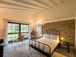 Un dormitorio con pared de piedra, una cama, una silla, una mesa y vista al jardín a través de un ventanal grande.