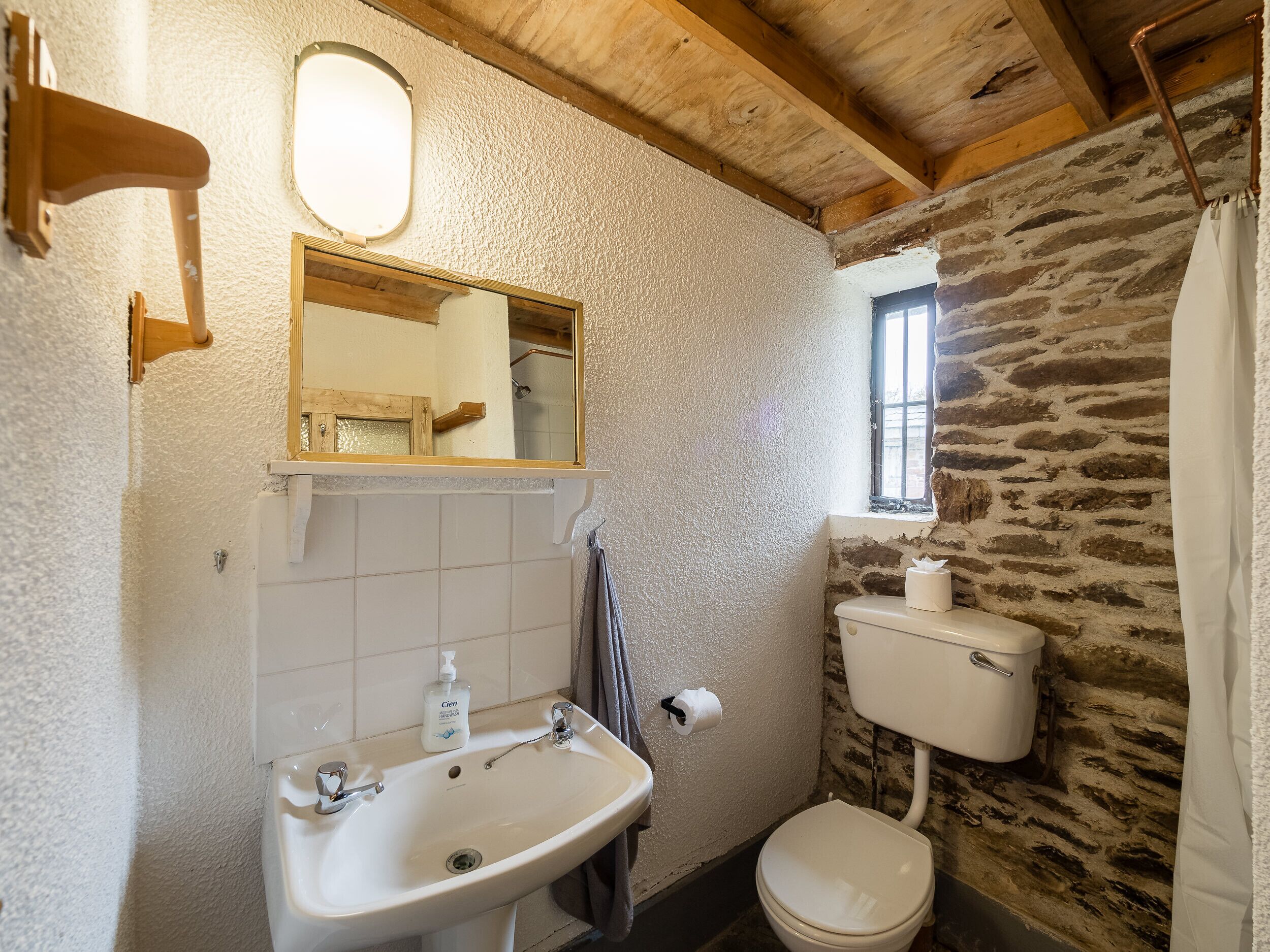 Een badkamer met een stenen muur, een houten plafond, een wit wastafelmeubel, een spiegel en een toilet.