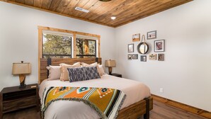 Un dormitorio con techo de madera, una cama grande con manta estampada, dos mesitas de noche con lámparas, y una ventana con vistas a árboles.