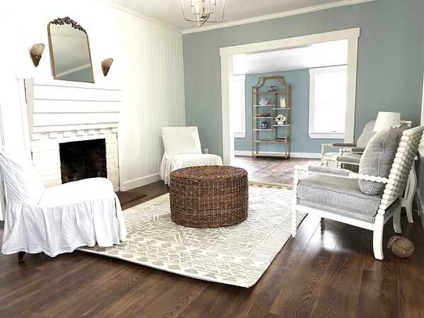 A living room with a fireplace, a white armchair, a grey sofa, a wicker ottoman, and a patterned rug.
