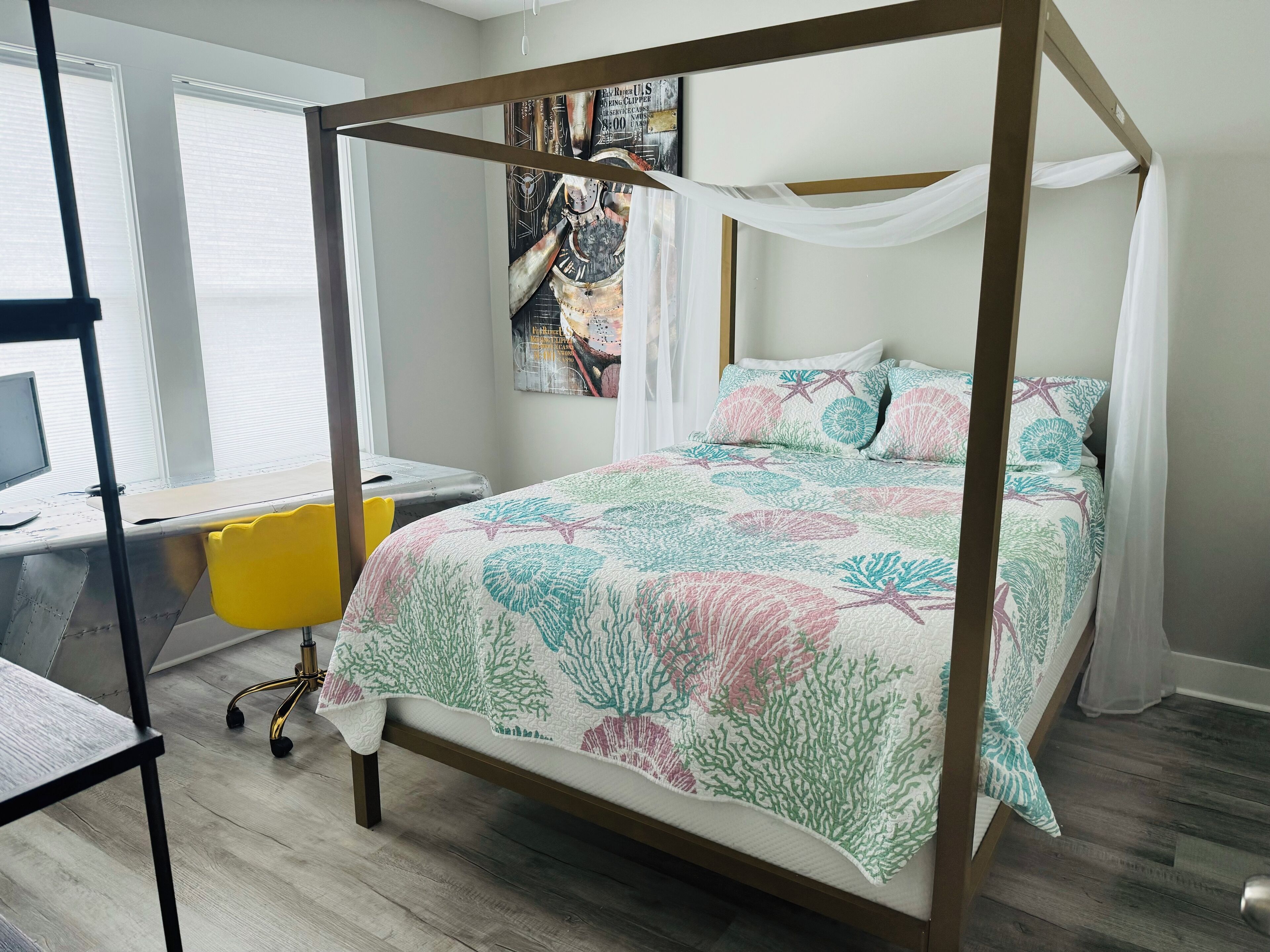 A four-poster bed with a floral bedspread, a desk with a yellow chair, and a computer monitor.