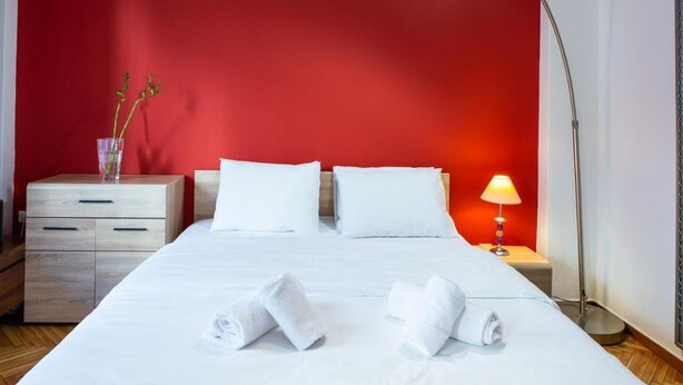 A hotel room with a red accent wall, a bed with white linens, two pillows, a bedside table with a lamp, and a nightstand with a vase of flowers. - Sleep in Style, Wake up in the Center of it All! (Thessaloniki)