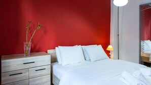 A hotel room with a bed, white bedding, a nightstand with drawers, a vase with green plants, a bedside lamp, and a mirror on the wall. - Sleep in Style, Wake up in the Center of it All! (Thessaloniki)