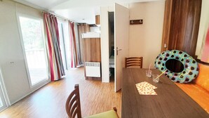 A compact room with a wooden dining table, a small chair, a colorful donut-shaped cushion, and a window with curtains. - Camping la Célestine (Beynes)