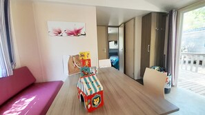 A room with a wooden floor, a pink sofa, a table with a colorful toy, and a bed in the background. - Camping la Célestine (Beynes)