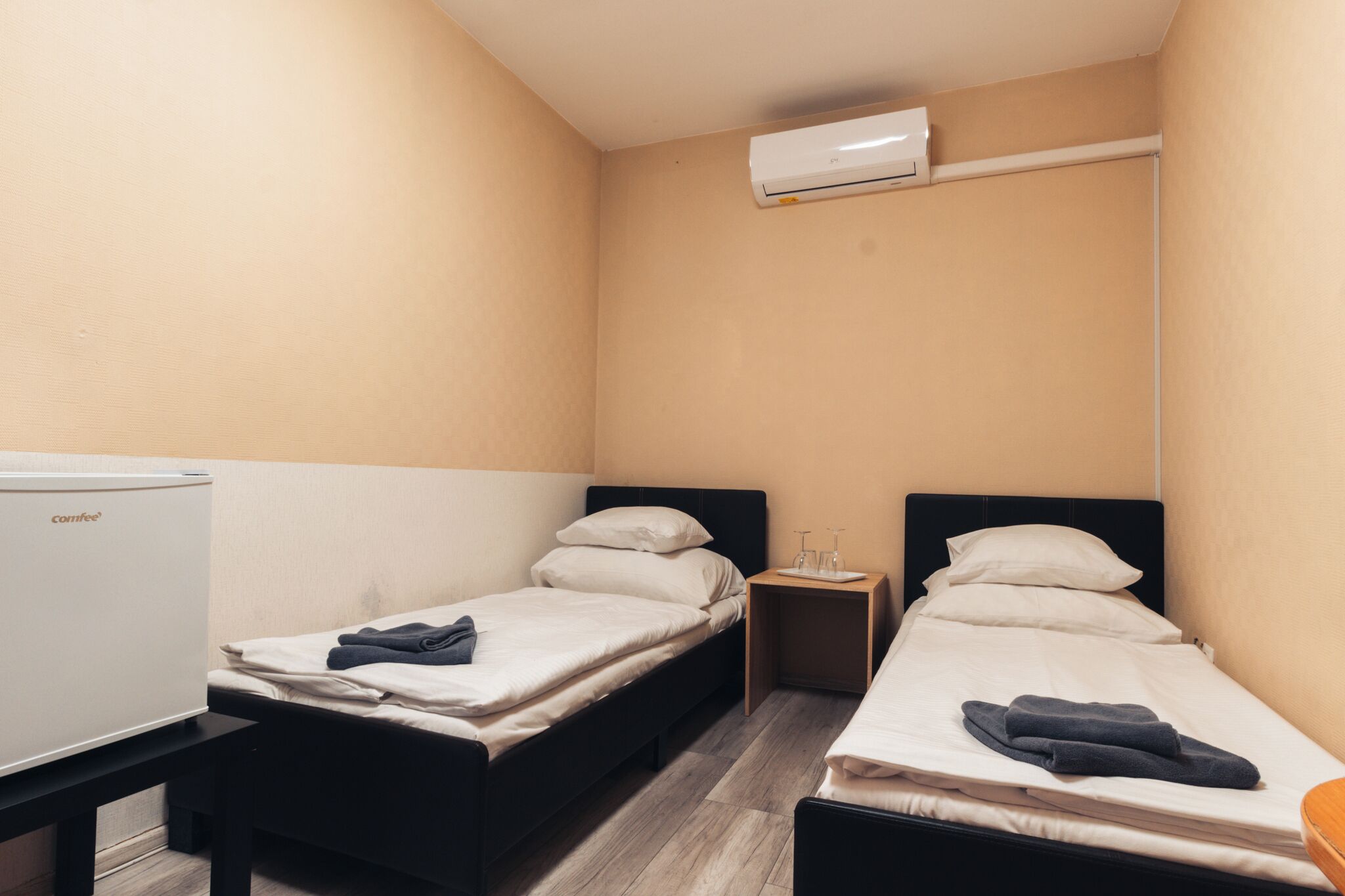 Een kleine hotelkamer met twee eenpersoonsbedden, een nachtkastje met een lamp, en een witte koelkast.