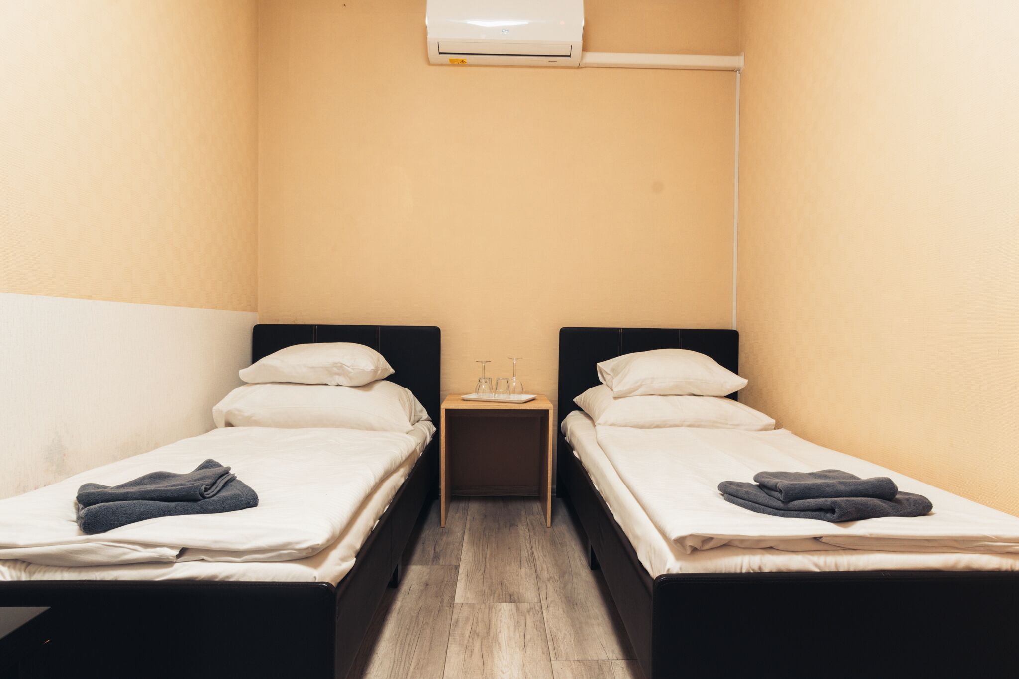 A small, clean room with two single beds, a bedside table with a lamp, and an air conditioning unit on the wall.