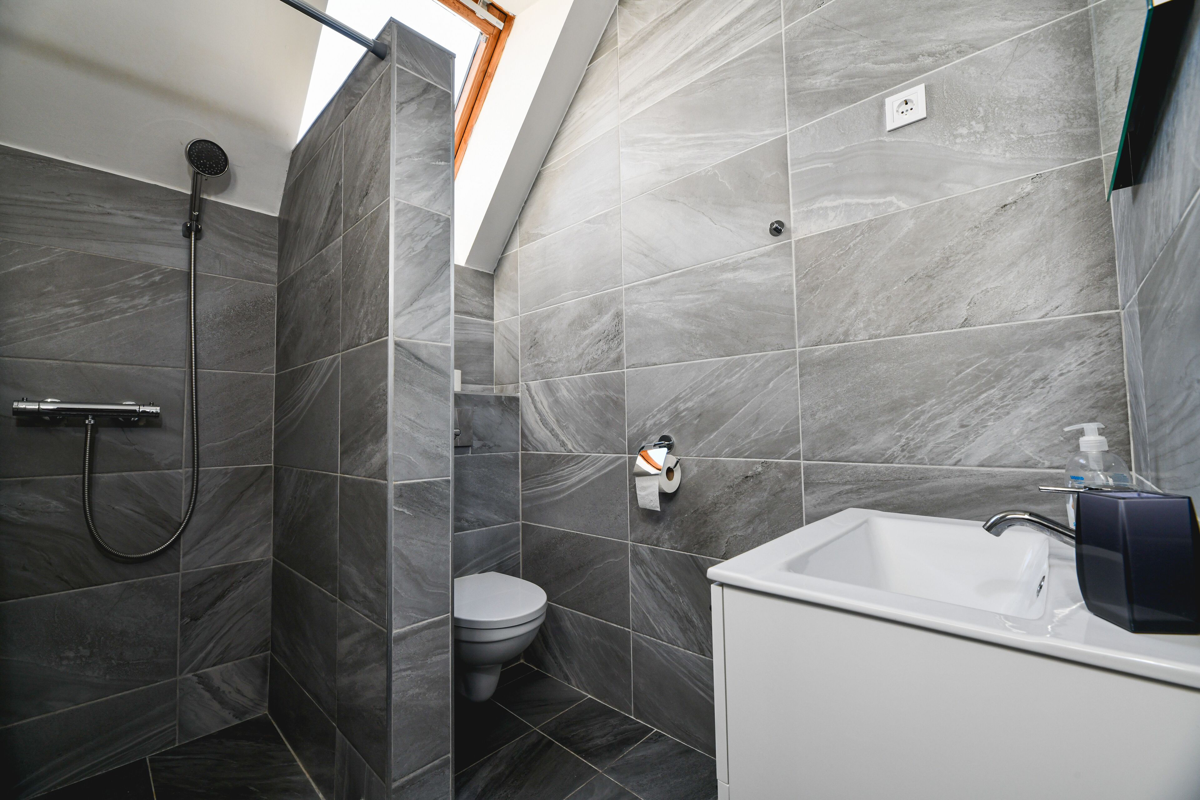 A modern bathroom with grey marble tiles, a shower, a toilet, and a white sink with a soap dispenser.