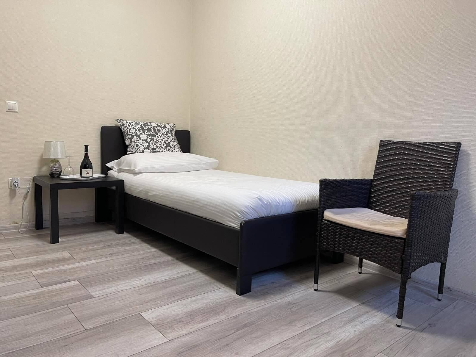 Un dormitorio pequeño y moderno con una cama individual, una mesita de noche con lámpara y una silla de mimbre.