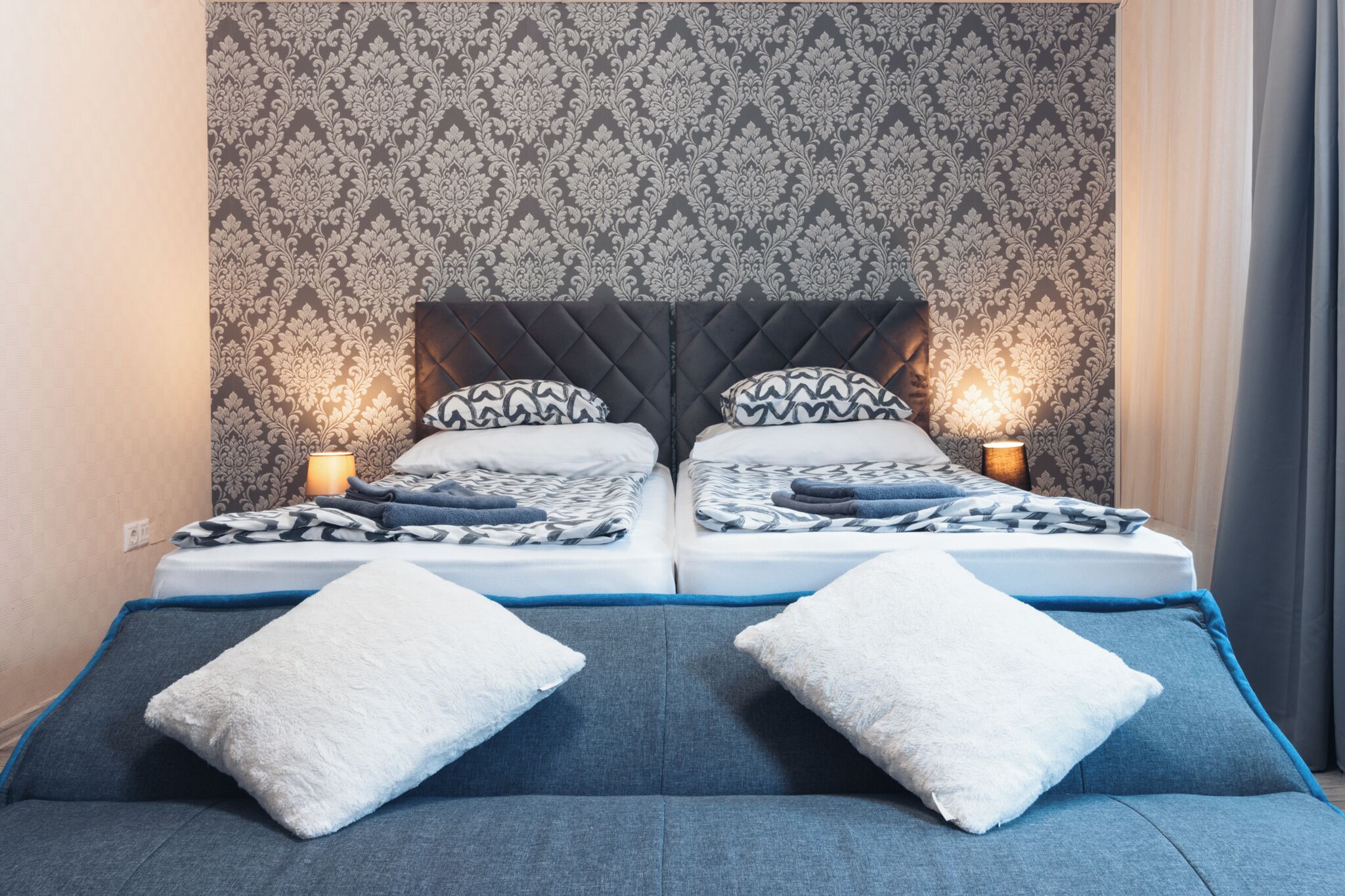 A double bed with a patterned headboard, two bedside tables with lamps, and a blue sofa with white cushions.