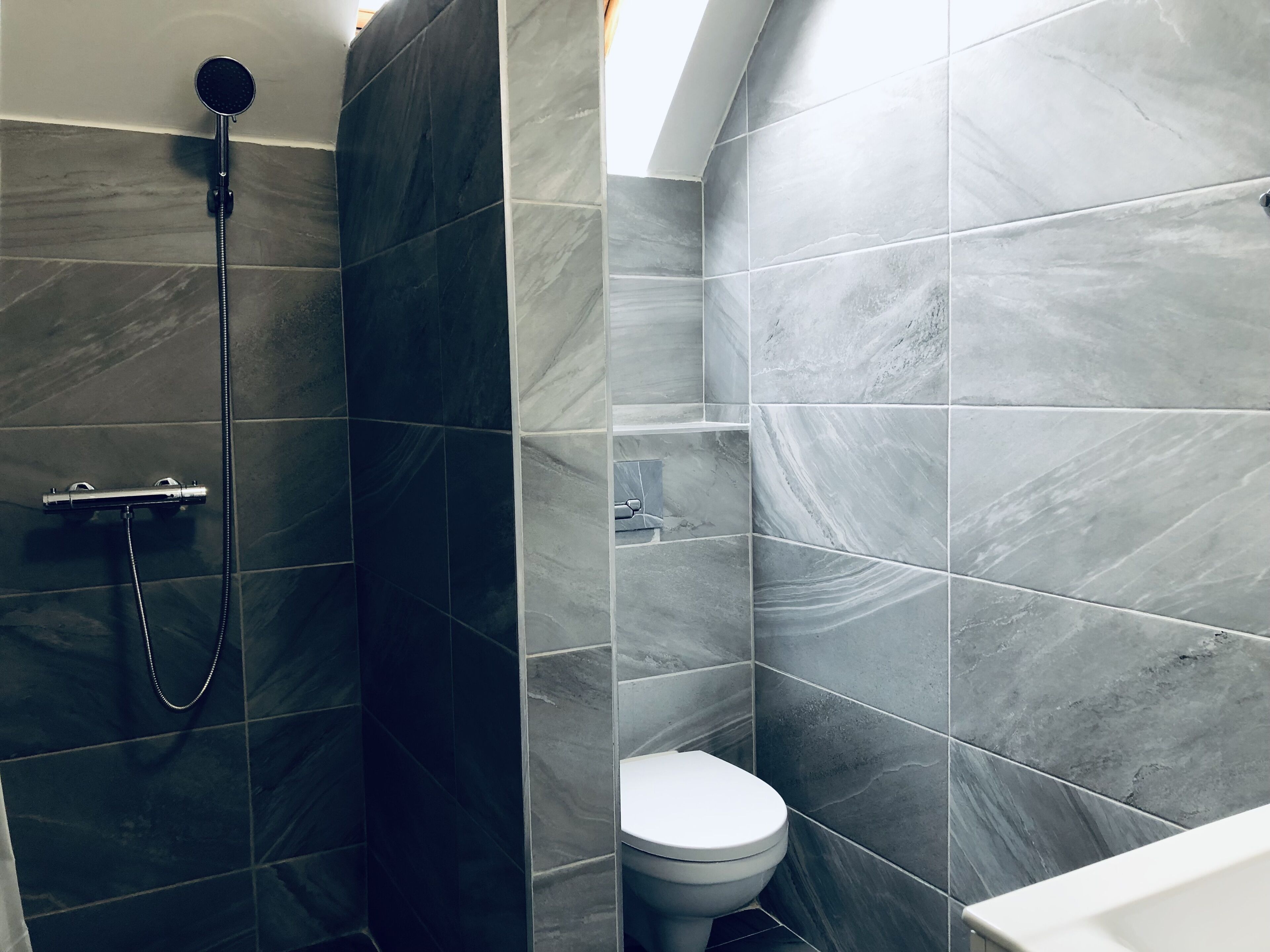 Un baño con baldosas gris oscuro, ducha con cabezal de mano y retrete.