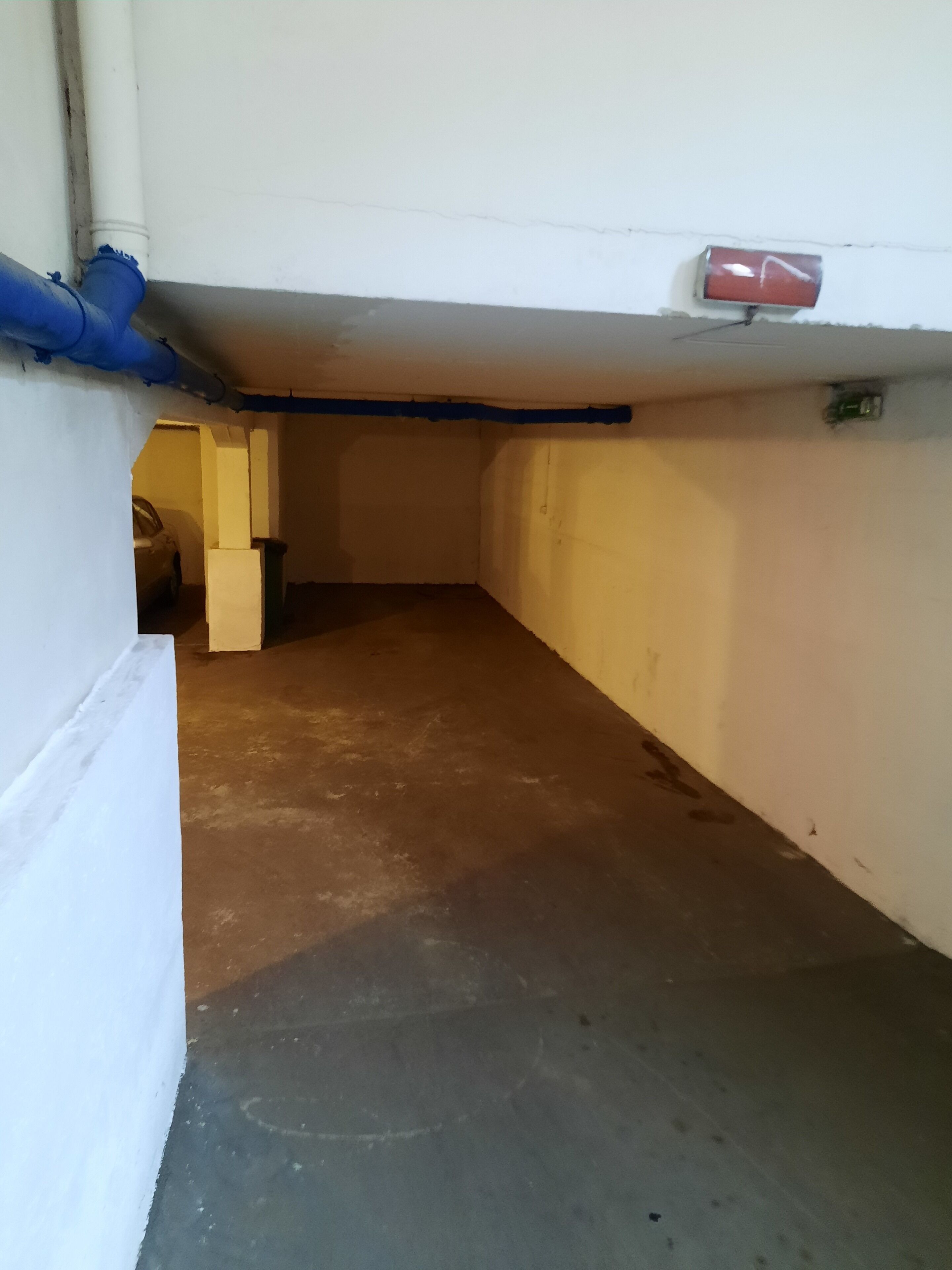 Een smalle gang met een betonnen vloer, witte muren en een blauwe buis die langs het plafond loopt.