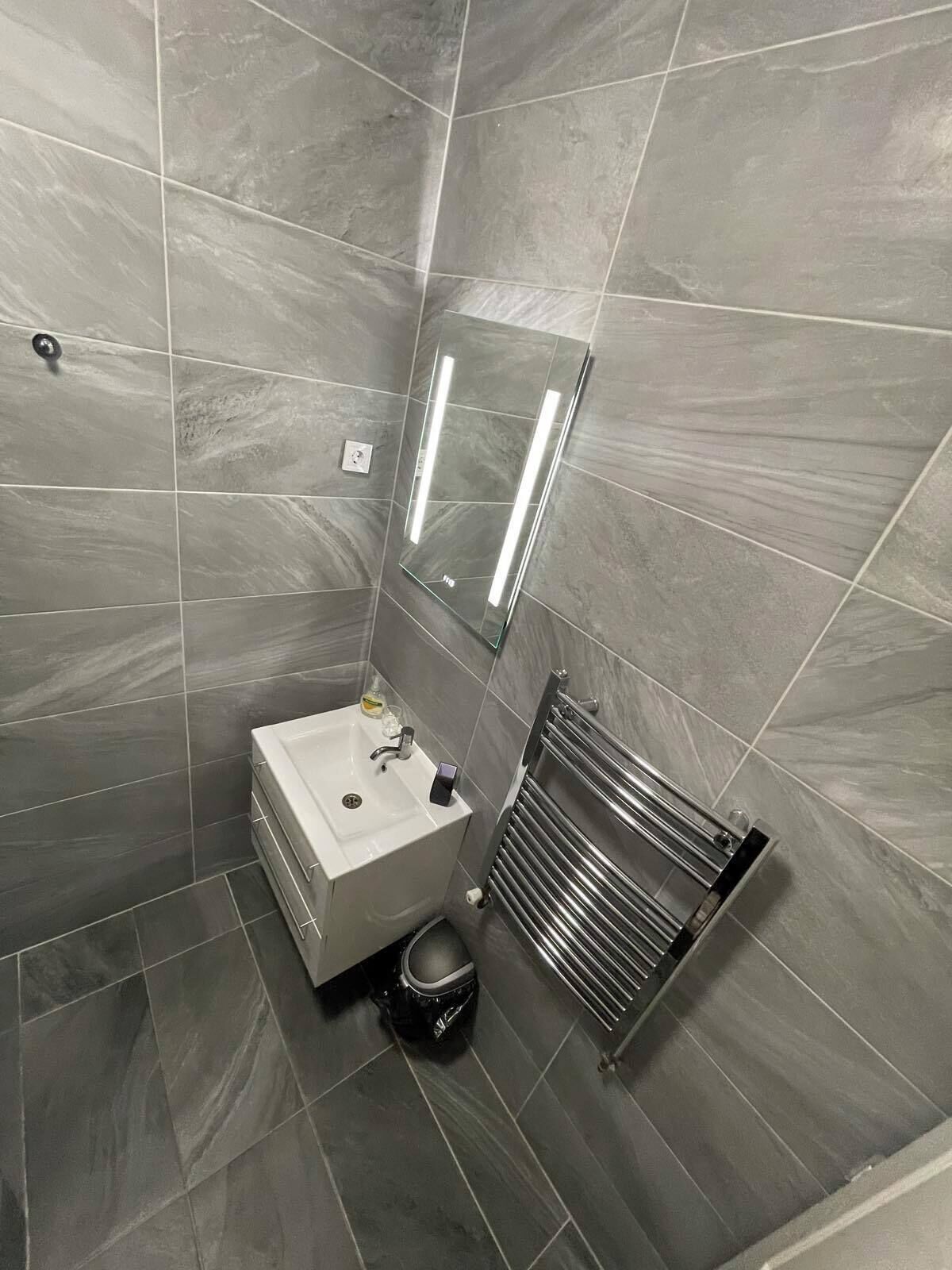 Casa de banho com azulejos cinzentos, uma sanita branca, um espelho e um porta-toalhas.