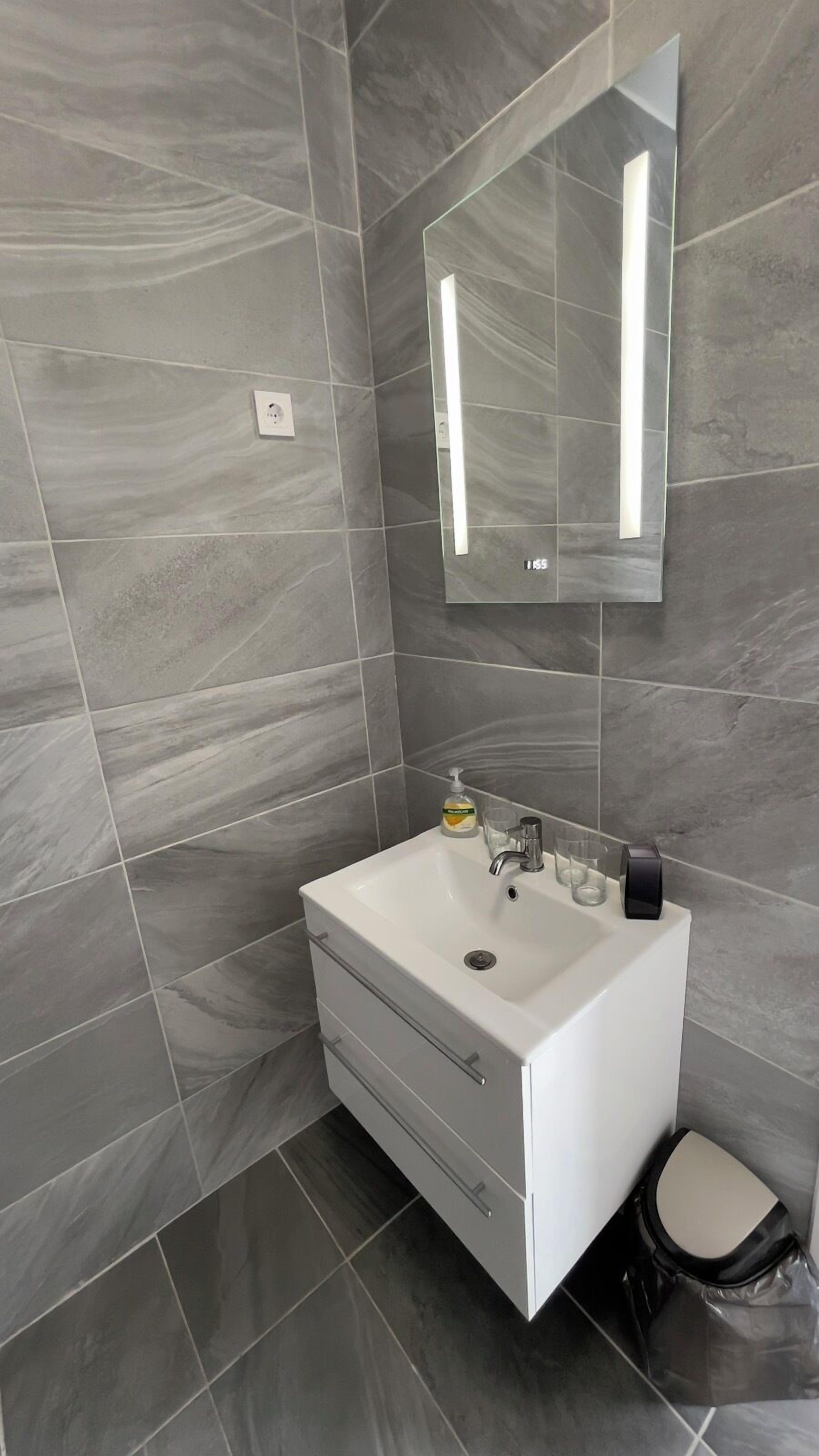 A modern bathroom with grey marble tiles, a white sink with a soap dispenser, a mirror, and a black trash bin.