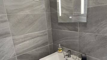 A modern bathroom with grey marble tiles, a white sink with a soap dispenser, a mirror, and a black trash bin.