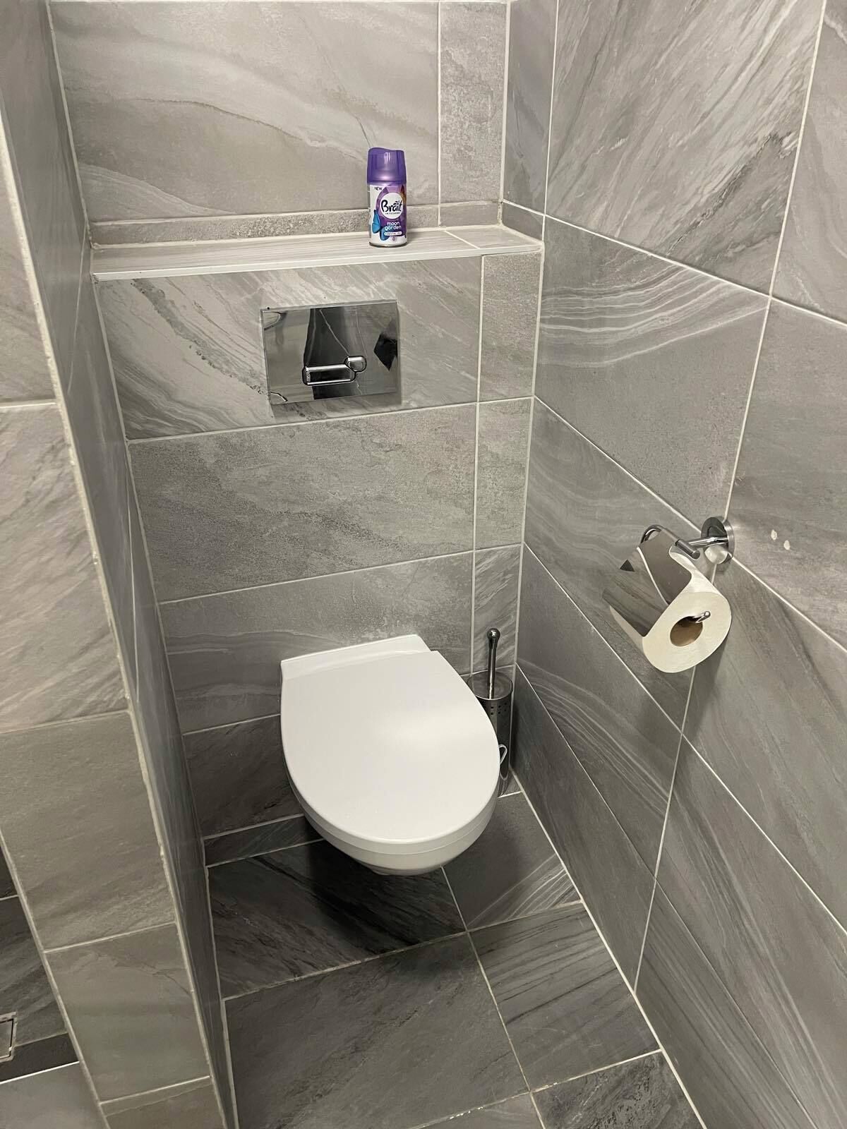 A small, modern bathroom with a white toilet, a shelf with a bottle, and a toilet paper holder.