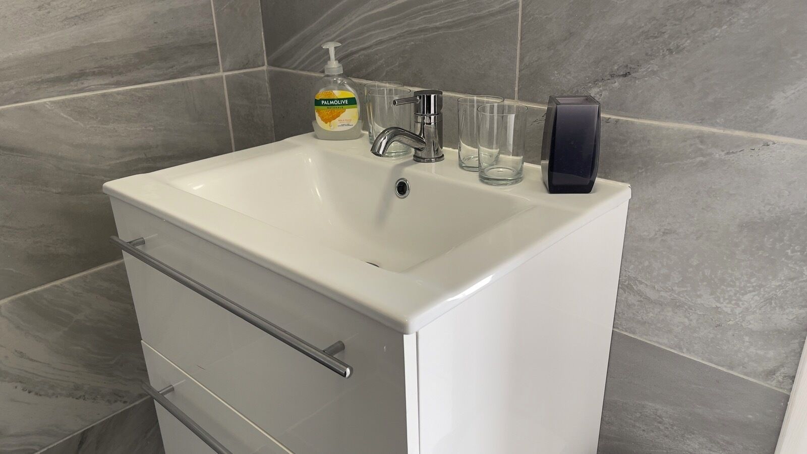 A bathroom with a white sink, a soap dispenser, and three clear glasses.