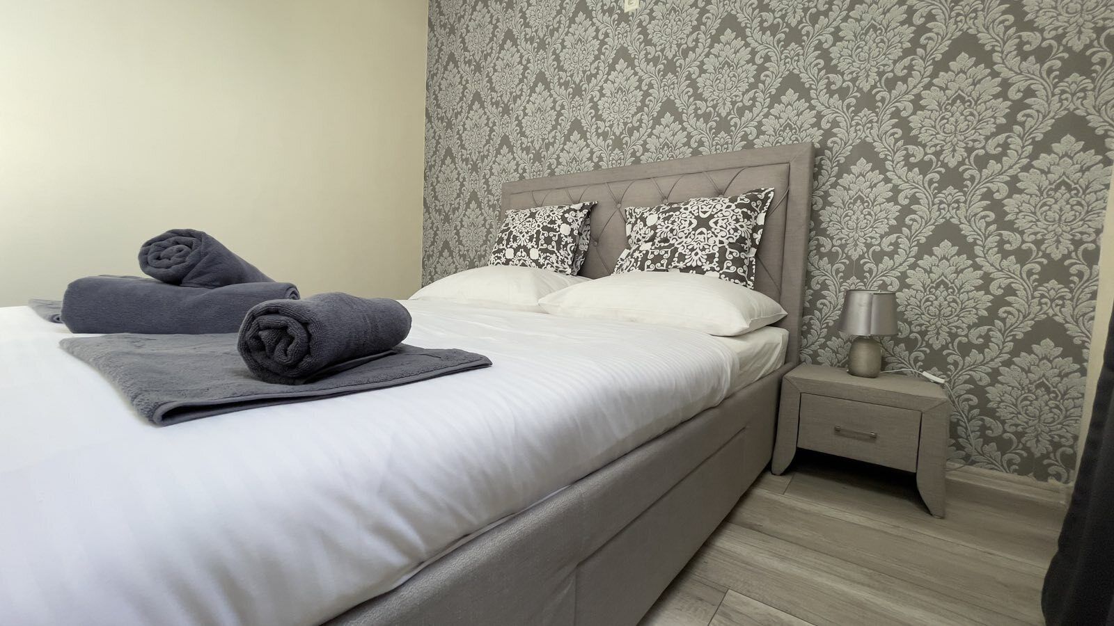 Una cama bien tendida con un cabecero gris, una mesita de noche con una lámpara y un papel tapiz estampado.