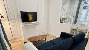 A living room with a dark blue sofa, a flat-screen TV mounted on a white wall, a small wooden table, and a view of a staircase and window overlooking a body of water. - Maison Amienoise Élégante (Amiens)